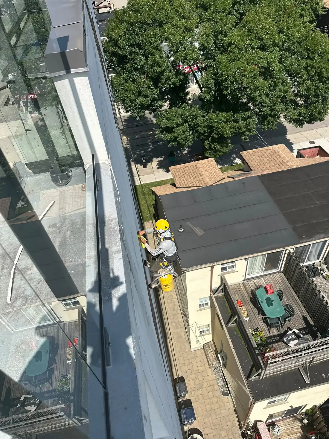 Rope access technician in helmet and full harness working a sealant joint on a high-rise wall, photographed from the roof above.