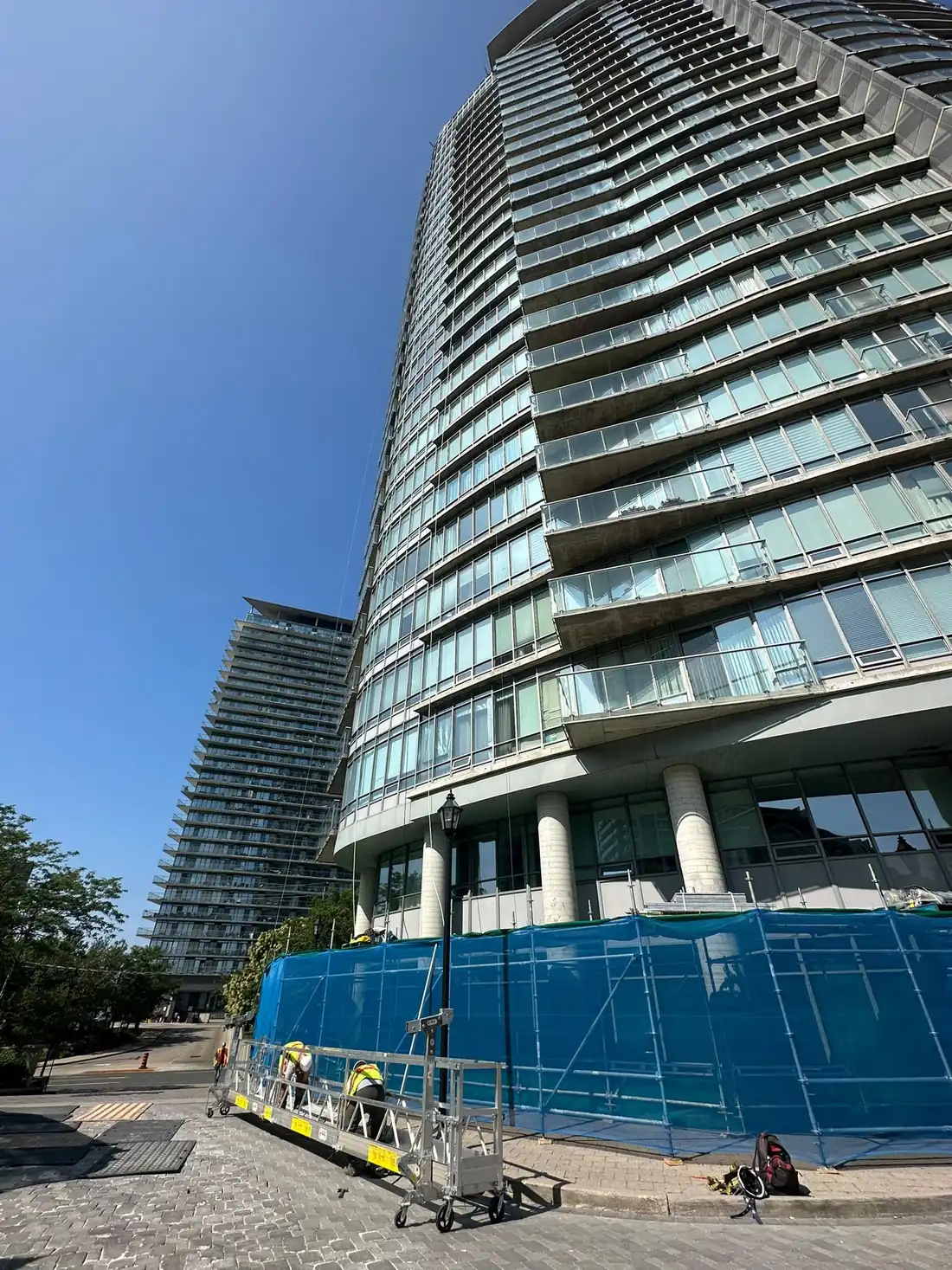 Swing stage rigged at the base of a curved condo tower with the crew in hi-vis, protective netting along the podium.