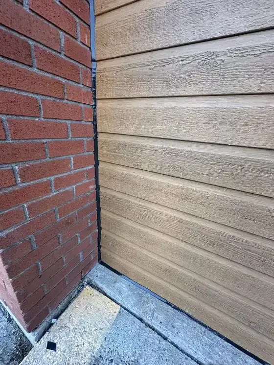 Fresh glossy black sealant bead running down a brick-to-siding transition and turning along the base at the concrete; the bead is neatly tooled and unmistakably the subject.
