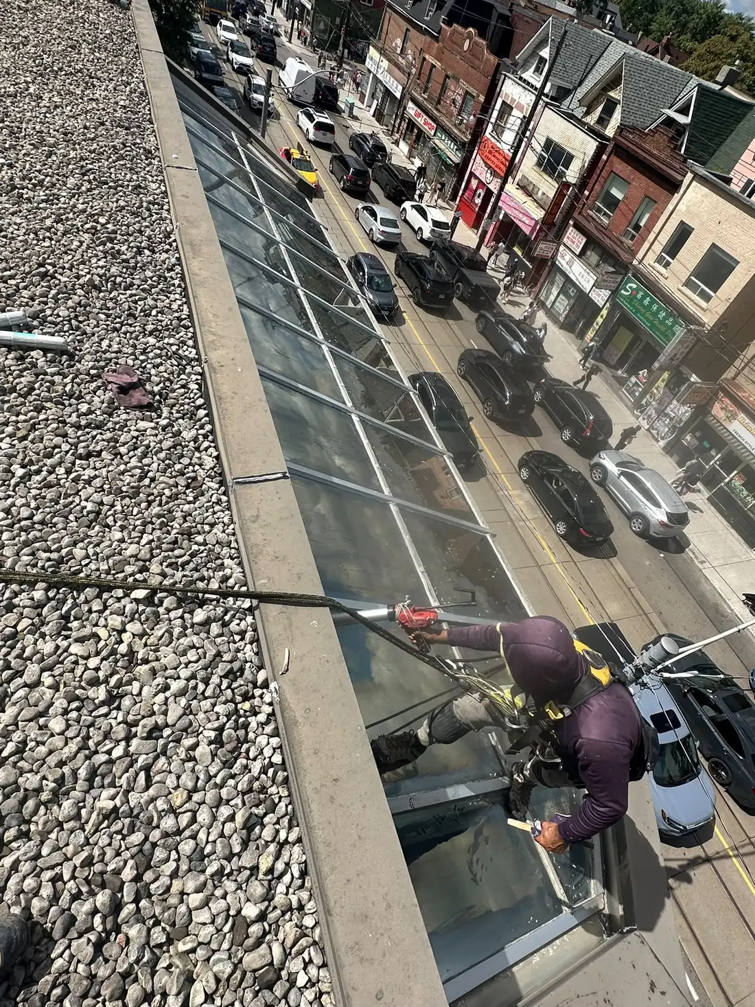 Rope access technician applying sealant with a caulking gun to a skylight glazing joint, working on ropes above a downtown street.