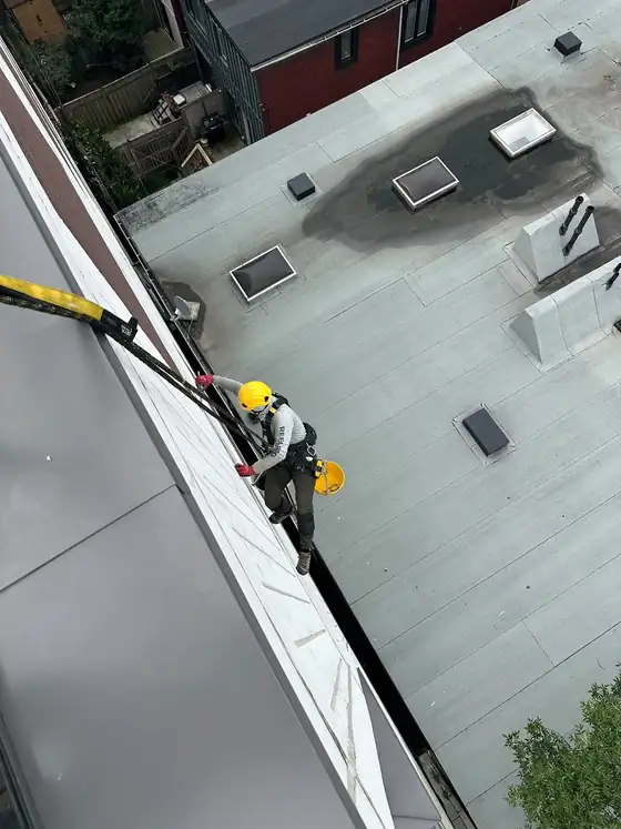 Technician on ropes at the parapet edge in helmet, harness and gloves, sealant bucket clipped to the harness.